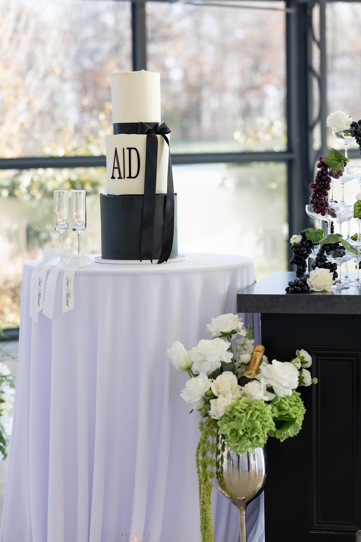 Three-tiered wedding cake on a white tablecloth with flowers and engraved champagne glasses in the background.