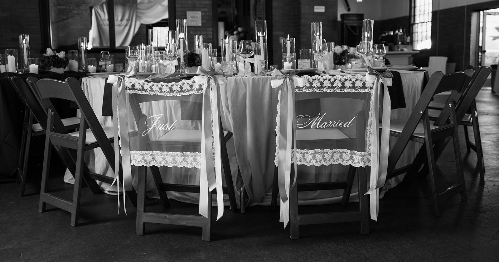Black and white setting of a wedding reception with chairs labeled 'Just Married' and a table with decorative items. Custom fabric wedding signage and bow details Kansas City